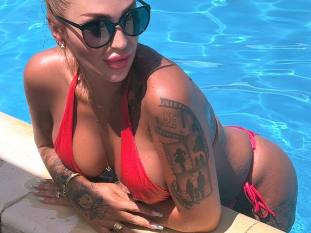 NicolSNaya en maillot de bain rouge, avec de grandes seins, des tatouages sur les bras et la main, portant des lunettes de soleil noires, appuyée sur le bord de la piscine, de l'eau en arrière-plan, peau bronzée, cheveux blonds..