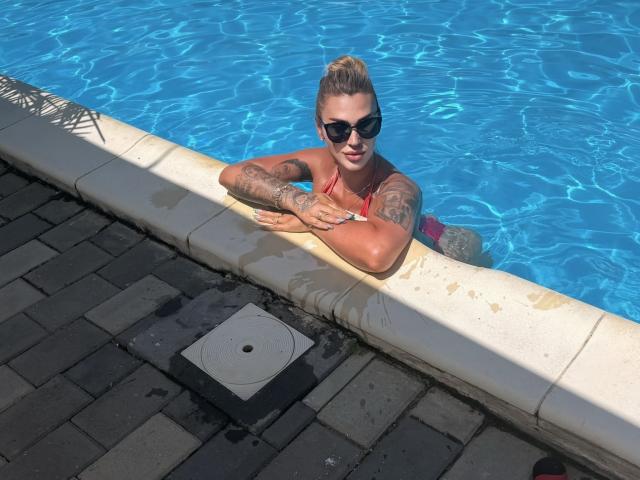 NicolSNaya avec des tatouages, les cheveux blonds attachés en chignon, des lunettes de soleil noires, un bikini rouge, les bras reposant sur le bord de la piscine, de l'eau bleue, un sol de piscine en briques grises, un bord de piscine blanc, souriante..
