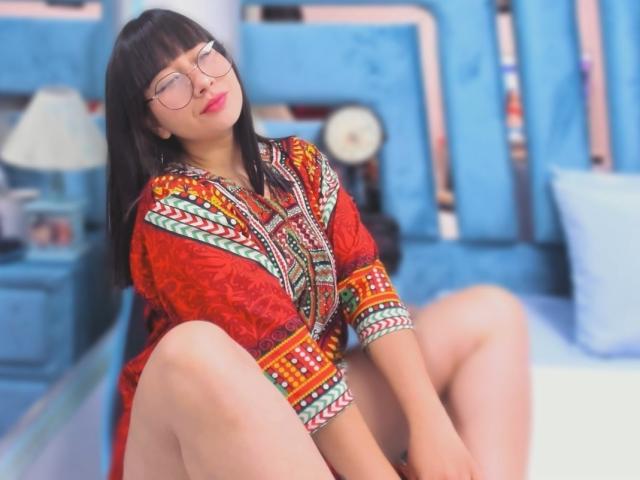 Genesiss with glasses, red and orange patterned dress, sitting with legs up, showing her bare ass, smiling, in a blue bedroom.