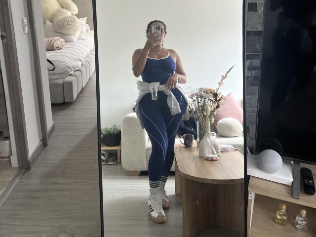 GalaMalkova with big tits, curvy ass, wearing a blue sports bra and matching pants, white sneakers, and a tied-up white shirt, taking a mirror selfie in a modern living room.