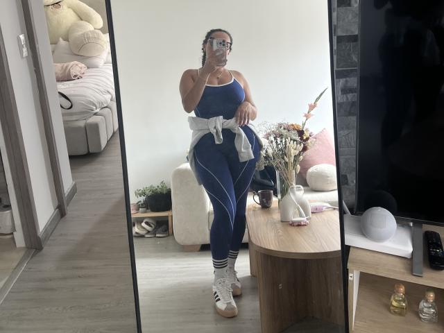 GalaMalkova, standing in front of a mirror, wearing a blue sports bra and matching pants, white socks with stripes, and Adidas sneakers, with a white shirt tied around her.