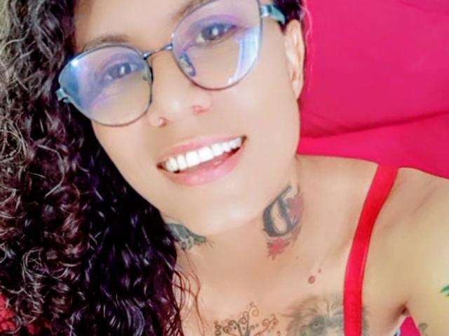 DamaLatina69, con cabello negro rizado, gafas azules, dientes blancos, un top rojo y tatuajes, sonriendo a la cámara. Sus pechos son pequeños y lleva gafas..