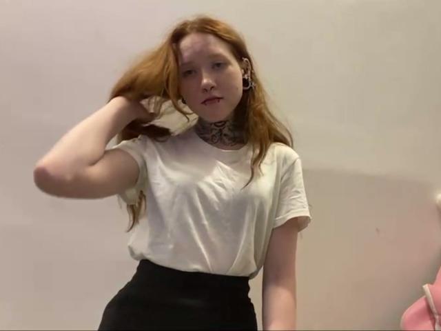 Machelle Trebesch ha la pelle chiara, i capelli rossi, una maglietta bianca, pantaloni neri a vita alta, un tatuaggio sul collo, piercing alle orecchie e tiene i capelli con una mano, apparendo leggermente annoiata..