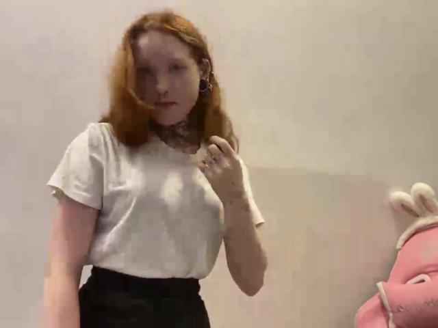 Machelle Trebesch, met rood haar, gekleed in een witte blouse en zwarte broek, staat met een hand in de buurt van haar borst en kijkt iets opzij. Een roze knuffeldiertje aan de rechterkant..