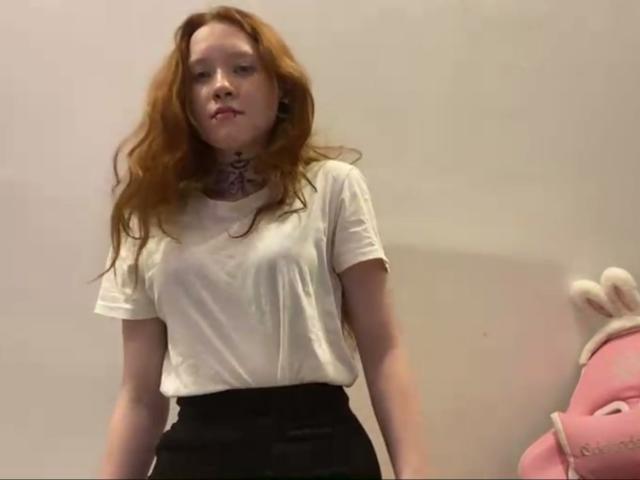 Machelle Trebesch, con cabello rojo ondulado, vestida con una camiseta blanca y pantalones negros de cintura alta, de pie con una ligera mueca, y un peluche rosa a la derecha..