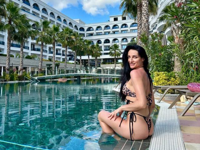TinaMart mit langen, schwarzen Haaren, die ein Bikini mit Leopardenmuster trägt, sitzt im Pool, zeigt ihr Gesäß, lächelt in die Kamera, im Hintergrund sind Palmen und tropische Pflanzen zu sehen..