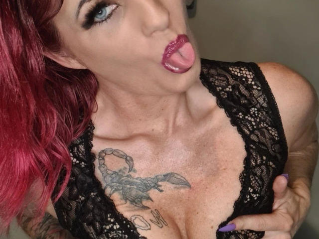 MiaMonez, avec des cheveux roux, un haut en dentelle noire, la langue tirée, du rouge à lèvres violet, une poitrine tatouée, une main sur la poitrine, des yeux bleus, du maquillage foncé et des ongles violets..