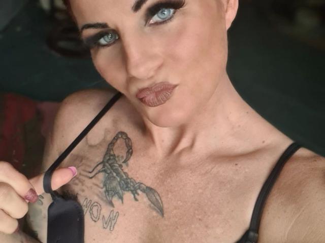 MiaMonez, aantrekkelijk met blauwe ogen, donkere make-up en een schorpioen-tatoeage op haar borst, houdt een zwarte band vast, met haar roze nagel wijzend naar de camera..