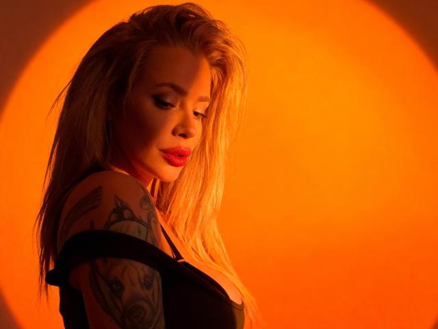 HotSadie, com cabelos loiros longos, batom vermelho e braço tatuado, vestindo uma blusa preta, iluminada por uma luz laranja, olhando para baixo com um sorriso discreto..
