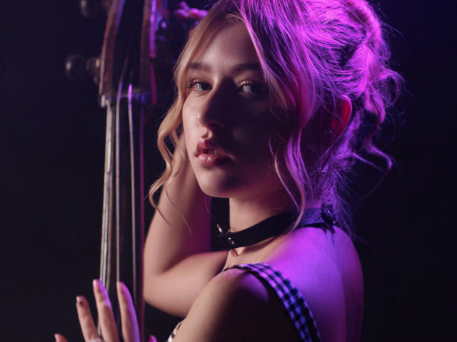Rosa Vance, con cabello rubio ondulado, reflejos rosados y un collar negro, sosteniendo un violín, mirando seductoramente a la cámara, con un vestido de cuadros que deja ver sus pequeños pechos..