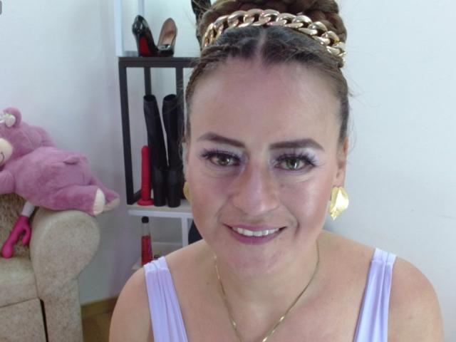 Maghentaa com cabelo castanho preso em um coque, tiara dourada, camiseta branca, sombra roxa, brincos dourados, sorrindo, porquinho de pelúcia rosa à esquerda, prateleira com sapatos ao fundo..
