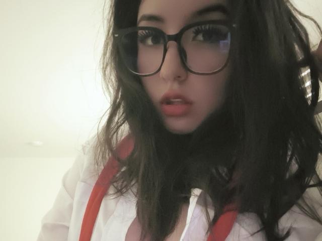 SusanMonroy with wavy dark hair, wearing red suspenders and a white shirt, has large glasses and slightly parted pink lips, showing her cleavage.