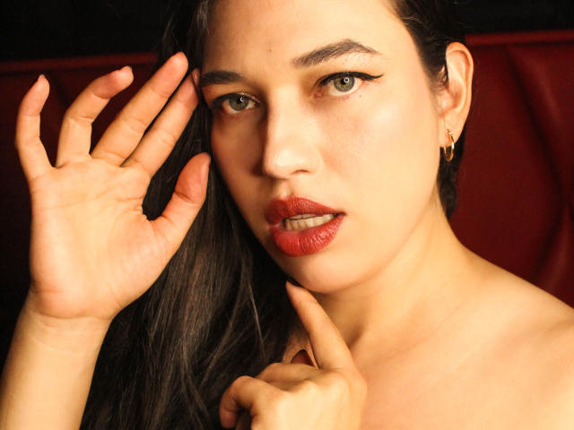 Rachel Wilson, com batom vermelho, sobrancelhas escuras, olhos verdes e pele brilhante, tocando o rosto e levantando a mão, exibindo seu nariz brilhante e lábios vermelhos levemente entreabertos..
