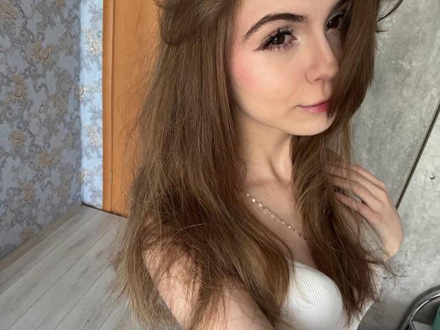 Lauralee Penderel, com cabelos castanhos longos, blusa branca sem alças, lábios rosados, sorriso discreto, mão perto do pescoço, olhos castanhos, pele clara, usando um colar fino, porta de madeira ao fundo..