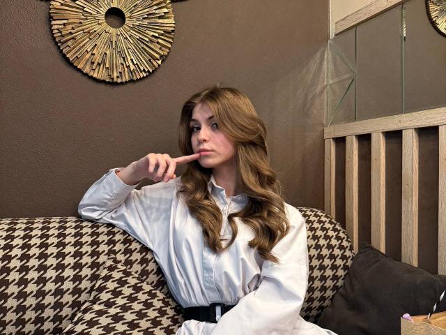 Lauralee Penderel, met lang, golvend bruin haar, gekleed in een witte blouse, zittend op een bank met een ruitpatroon, met haar vinger in haar mond, en met een zwarte riem..