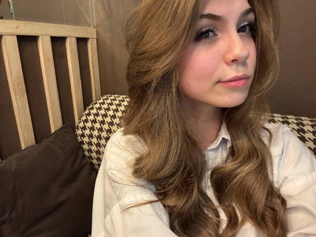 Lauralee Penderel, met lang, golvend bruin haar, een witte blouse en subtiele make-up, toont een lichte glimlach en zit op een kussen met een ruitpatroon, met een houten hoofdeinde op de achtergrond..