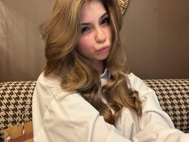 Lauralee Penderel, met golvend blond haar, volle roze lippen en een witte blouse, zit op een bank met een ruitpatroon en kijkt naar de camera. Haar borsten zijn klein en ze is schattig..
