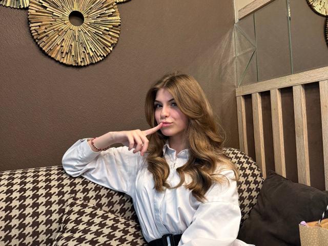 Lauralee Penderel, con el pelo largo y ondulado de color marrón, una camisa blanca abotonada y un dedo en sus labios rosados, sentada en un sofá con estampado de cuadros de galgo, sonriendo ligeramente..