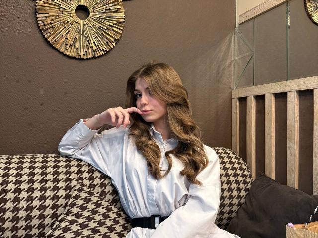 Lauralee Penderel, met lang, golvend bruin haar, draagt een witte blouse en zit op een bank met een ruitpatroon. Ze heeft haar vinger op haar lippen en glimlacht licht. Haar achterkant is gedeeltelijk zichtbaar..