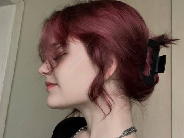 NenaSelic, avec des cheveux rouge-rose attachés en un chignon désordonné, une pince à cheveux noire, un anneau de nez et un collier en chaîne, les yeux fermés, tournée vers la gauche. Peau pâle, sourire discret..
