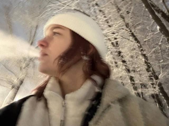 Nena Selic, met een witte gebreide muts, die een wolkje uitblaast, draagt een witte hoodie en staat in een besneeuwd bos, waarbij ze met haar bruine haar naar de zijkant kijkt..