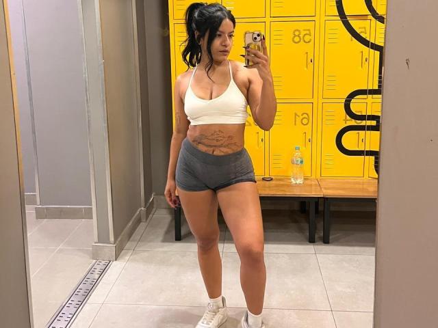 Sophie Soler met grote borsten, korte grijze broekjes, een witte sportbeha en witte sneakers, die een telefoon vasthoudt en staat in een kleedkamer met gele kluisjes..