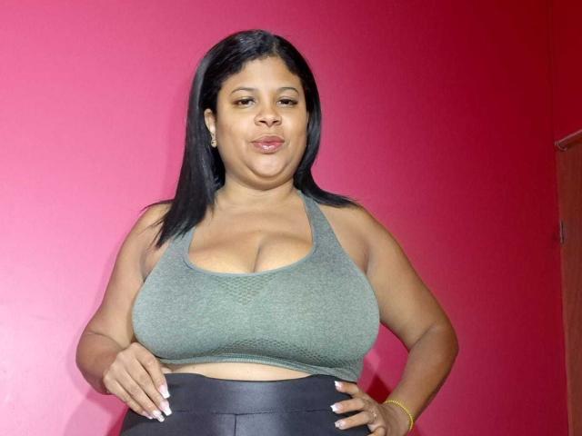 NaughtyPlusSizeGoddess met donkere huid, zwart haar, een groene sportbeha, waarbij haar grote borsten zichtbaar zijn, zwarte broek, een roze muur, een gouden armband en een lichte frons. Handen op de heupen, met een zelfverzekerde blik..
