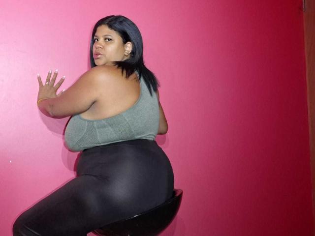 NaughtyPlusSizeGoddess met grote borsten, zwart haar, een groene top, zwarte broek, zittend op een zwarte kruk, tegen een roze muur, kijkt om, rechterhand op de muur, lichte frons..