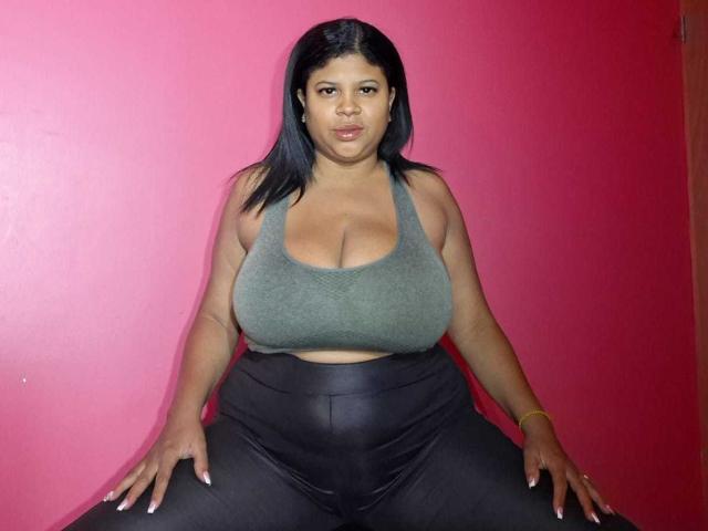 NaughtyPlusSizeGoddess mit großen Brüsten in einem grünen Sport-BH, schwarzen Lederhosen, sitzend mit gespreizten Beinen, schwarzes Haar, braune Augen, rosa Hintergrund, gelanges Armband, selbstbewusster Ausdruck..