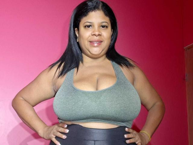 NaughtyPlusSizeGoddess con cabello oscuro, sostén deportivo verde, mostrando grandes senos, manos en las caderas, pantalones negros, pulsera amarilla, sonriendo, pared rosa de fondo..