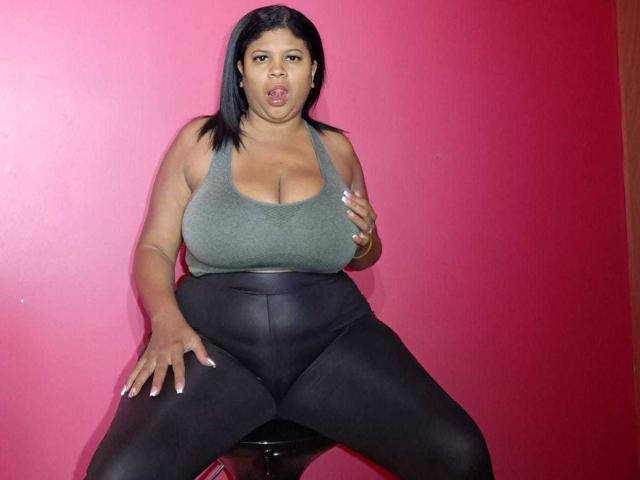 NaughtyPlusSizeGoddess con grandi seni, vestita con una canotta verde, leggings neri e capelli neri, seduta su uno sgabello contro un muro rosso, con la bocca aperta..