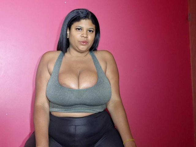 NaughtyPlusSizeGoddess has big boobs in a gray sports bra, black leather pants, and black hair, sitting against a pink wall, looking at the camera with a neutral expression.