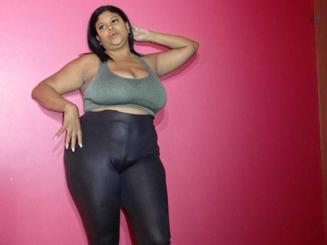 NaughtyPlusSizeGoddess con piel marrón media, cabello negro, un top deportivo verde y pantalones de cuero negro, de pie frente a una pared rosa, con una mano en la cabeza, mostrando senos grandes y brillantes..