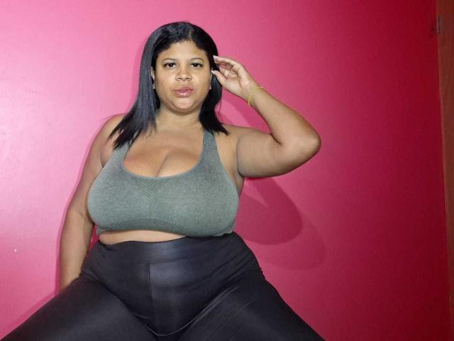 NaughtyPlusSizeGoddess avec une peau brun moyen, des cheveux noirs et lisses, un soutien-gorge de sport vert, montrant de grandes seins, un pantalon noir taille haute, la main droite dans les cheveux, devant un mur rouge..