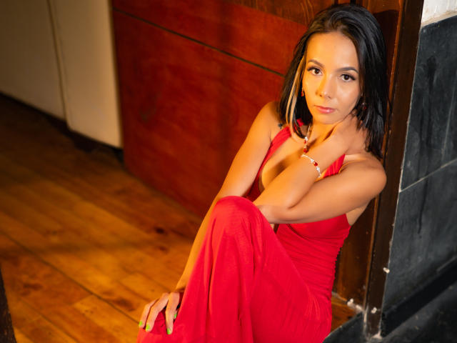 EvelinLuna, avec de petites seins, assise sur un plancher en bois, portant une robe rouge, cheveux noirs, ongles verts, mur en bois, carrelage noir, regardant vers le haut, main sur la poitrine, sourire discret..