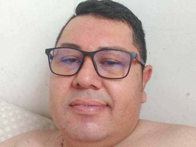 TheCuteCrazy, ein etwas korpulenter Mann mit kurzen, schwarzen Haaren, der eine schwarze Brille trägt, hat violett schimmernde Augen und ein leichtes Lächeln, ist ohne Hemd vor einer weißen Wand..