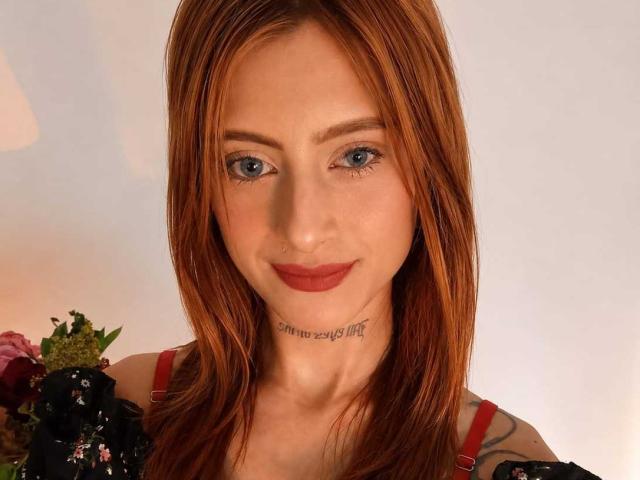 EliMonroe hat rotes Haar, blaue Augen und ein Tattoo unter ihrem Kinn. Sie trägt ein rotes Kleid mit Trägern und hat einen kleinen Nasenring. Ihre Brüste sind nicht sichtbar..