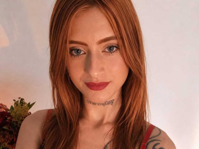 EliMonroe hat rotes Haar, blaue Augen und ein Tattoo unter ihrem Kinn. Sie trägt roten Lippenstift und ein rotes Oberteil, und hat ein Tattoo auf ihrem linken Arm..