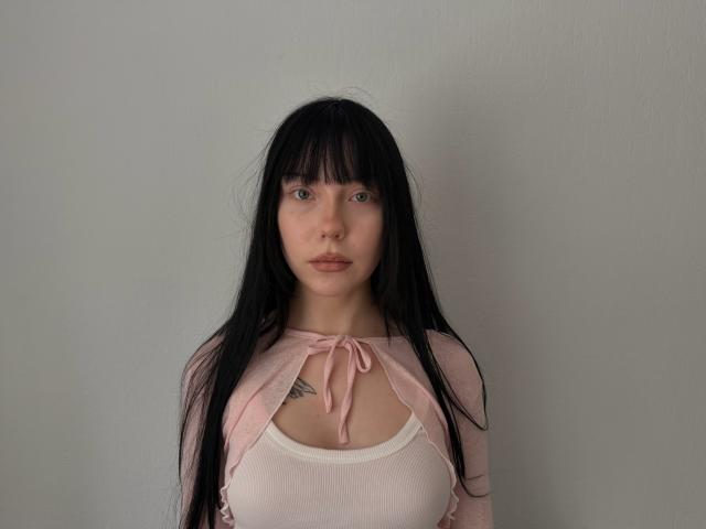 EllenChristoph, met lang zwart haar, een lichtroze vest en een witte top, toont kleine borsten en een tatoeage onder haar borst, heeft een neutrale uitdrukking en staat tegen een effen muur..