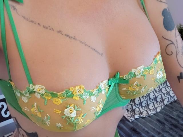 French : Shanna porte un soutien-gorge floral vert et jaune qui met en valeur sa poitrine, dévoilant sa poitrine et des tatouages sur son bras droit. Sa peau est claire, et elle porte une bretelle verte..