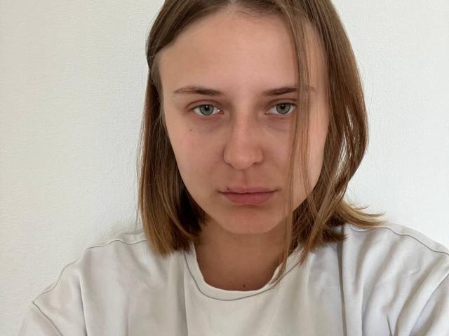 Lera Kreiger hat glattes, braunes Haar, grüne Augen und einen neutralen Gesichtsausdruck. Sie trägt ein weißes Hemd und blickt direkt in die Kamera, mit einem leicht schmollenden, entspannten Gesicht..