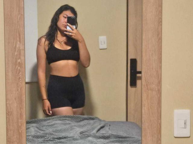 ValeryMartinez has medium brown skin, black wavy hair, black sports bra, and black shorts, standing in front of a mirror, taking a selfie with her phone.