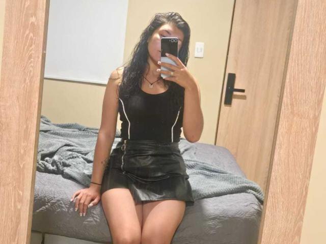 ValeryMartinez with wet black hair, wearing a black tank top and black leather skirt, sitting on a bed, taking a mirror selfie with her phone, showing her ass.