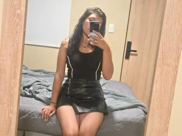 Valery Martinez, con i capelli neri bagnati, indossa una canotta nera e una gonna in pelle, seduta su un letto, sta scattando un selfie allo specchio, mostrando le sue cosce nude e il suo piccolo sedere..