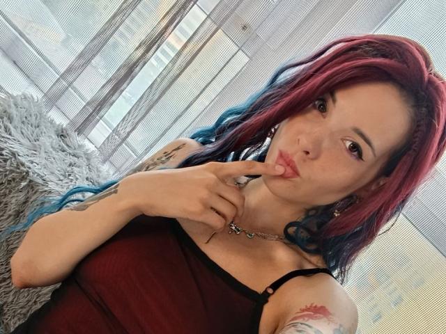RachelNervis, avec des cheveux roux et bleus, des lèvres pulpeuses, touchant ses doigts, portant un débardeur rouge, montrant des tatouages, regardant la caméra, assise sur un coussin gris et duveteux..