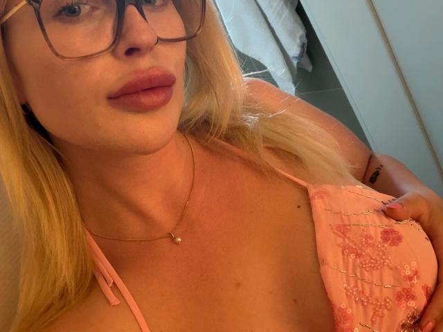 Hellybunny-hot with blonde hair, glasses, pink floral top, showing her tits, a gold necklace, and pink lipstick. Her hand's on her chest. Her nipples aren't visible.