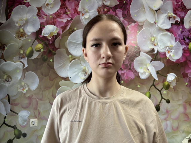 PastyAylsworth, a young woman with pale skin and brown hair in a ponytail, wearing a beige shirt, standing in front of a wall of white and pink orchids.