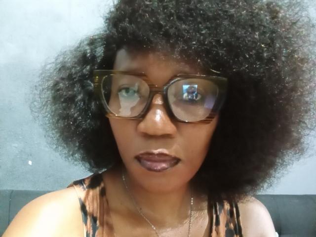 TeeGlam, con cabello rizado oscuro, gafas marrones estilo carey y lápiz labial morado, vistiendo una blusa estampada, mirando directamente a la cámara con una expresión seria..