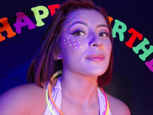 LizBonnet mit neonfarbenen Punkten auf dem Gesicht, pinkem Lippenstift und braunen Haaren, trägt ein farbenfrohes Halstopp und hat ein selbstbewusstes Lächeln sowie große Ohrringe, vor einem dunkelblauen Hintergrund..