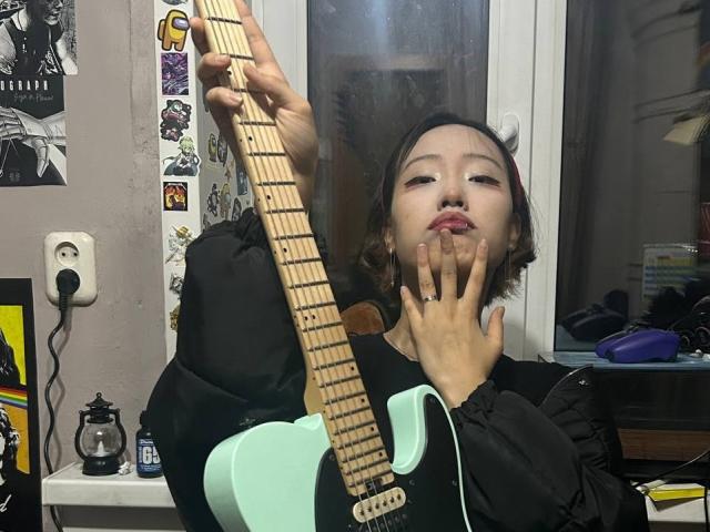Odette Arenstam che suona una chitarra elettrica color verde menta, con le labbra gonfie, le mani vicine al viso, indossa una giacca nera e ha un piercing al naso, in una stanza poco illuminata..
